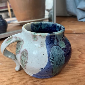 Könnte beinhalten: Handgefertigte Keramiktasse mit rundem Körper und gebogenem Henkel. Die Tasse hat eine weiße Basis mit blau, grün und violett glasierter Oberfläche. Der Innenraum ist tiefblau. Die Tasse steht auf einer Holzoberfläche.