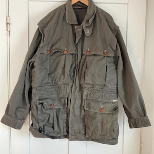 May include: Olive green jacket with multiple front pockets and wooden buttons. The jacket has a collared neckline and is hanging on a wooden hanger. The jacket appears to be made of a durable, possibly canvas-like material.