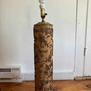 May include: A tall, cylindrical floor lamp with a beige, textured shade featuring a floral design. The lamp has brass accents at the top and base, and a standard light bulb is screwed into the socket. The lamp stands approximately 50 inches tall.