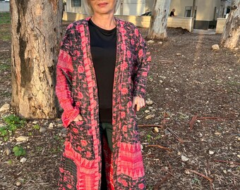 Pode incluir: Um casaco comprido e aberto com um padrão floral vibrante em tons de vermelho e preto. O casaco apresenta um design de patchwork e bolsos. A pessoa está a usar óculos de sol, uma camisa preta, calças verdes e ténis brancos.