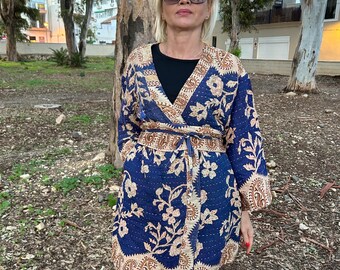 Pode incluir: Uma jaqueta azul estilo quimono com um padrão floral em bege e branco. A jaqueta tem um design envolvente com um cinto a condizer. As mangas são compridas e a jaqueta é usada sobre uma blusa preta e calças verdes.
