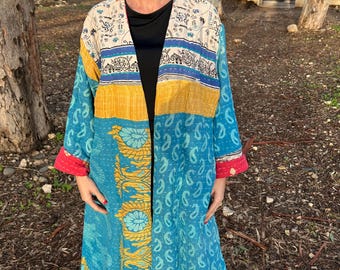 Pode incluir: Um kimono longo e aberto na frente em tons de azul, amarelo e creme. O kimono apresenta um padrão paisley e floral, com detalhes em vermelho nos punhos. A roupa é usada ao ar livre.