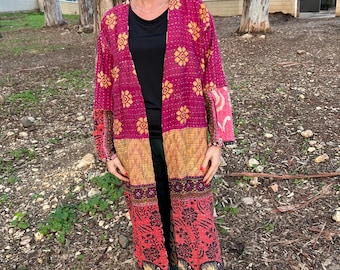 Pode incluir: Um longo quimono magenta com frente aberta e um padrão floral em dourado e vermelho. O quimono tem um design de patchwork com painéis contrastantes de diferentes padrões e cores. As mangas e a bainha apresentam detalhes intrincados, usado sobre uma blusa preta e calças verdes.