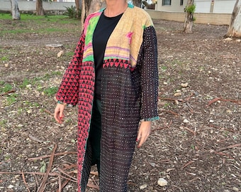 Pode incluir: Um casaco comprido estilo quimono em patchwork com uma variedade de cores e padrões. O casaco apresenta uma base preta com pontos brancos, triângulos vermelhos e detalhes florais. O casaco é usado sobre um top preto e calças verdes.