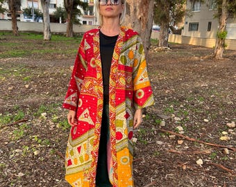 Pode incluir: Um kimono longo e aberto na frente em vermelho, amarelo e verde vibrantes. O kimono apresenta um padrão paisley e geométrico. A pessoa está usando um top preto, calças verdes e tênis brancos. O fundo inclui árvores e um edifício.