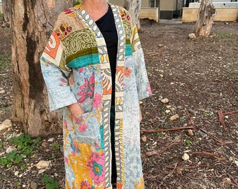Pode incluir: Robe longa estilo quimono em patchwork com uma variedade de padrões florais e geométricos em tons de azul, amarelo, rosa e verde. O robe tem mangas compridas e bolsos. A pessoa está usando óculos de sol e tênis brancos.