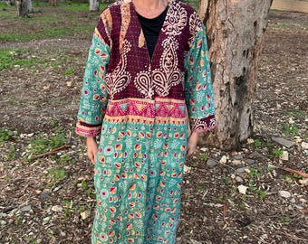 Pode incluir: Um casaco comprido estampado em tons de bordô, verde-azulado e rosa. O casaco apresenta um design floral detalhado e um fecho de correr. A pessoa está usando tênis brancos e óculos de sol escuros.