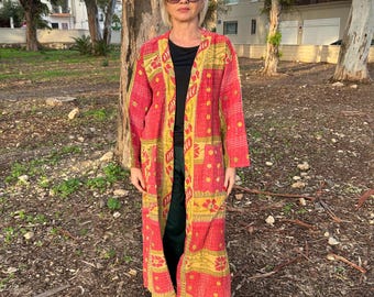 Pode incluir: Casaco comprido estilo quimono, aberto, com padrão vermelho e amarelo. O casaco apresenta um design floral e geométrico repetitivo. A pessoa está a usar um top preto, calças verdes e ténis brancos. O fundo inclui árvores e um edifício.