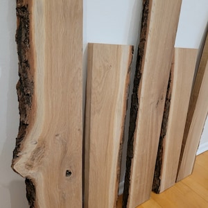 Könnte beinhalten: Mehrere unbearbeitete, hellfarbene Holzbretter mit naturbelassenen Kanten lehnen an einer weißen Wand. Die Bretter variieren in Breite und Länge und zeigen die natürliche Maserung und Textur des Holzes. Die Kanten haben eine dunkle, raue Rinde.
