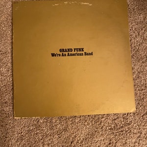 May include: A gold vinyl record album with the text "GRAND FUNK We're An American Band" printed in black.