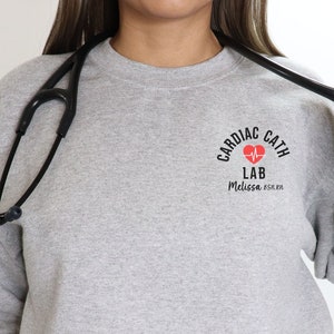 May include: Gray sweatshirt with a black and red graphic that says "Cardiac Cath Lab" with a heart and EKG line. The text "Melissa Asken" is below the graphic.