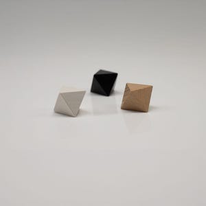 May include: Three geometric octahedron-shaped objects arranged on a surface. One object is light gray, one is solid black, and one is a natural light brown wood grain. Each piece features multiple triangular faces meeting at a central point.