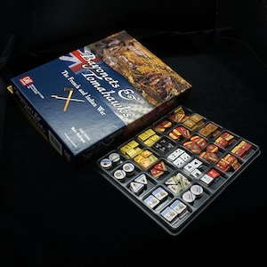 以下が含まれることがあります： 青と赤のカバーが付いたボードゲームの箱で、「バヨネット＆トマホーク：フランスとインディアンの戦争」という文字が書かれています。箱が開いており、木製のサイコロ、プラスチック製のトークン、厚紙製のカードなど、ゲームの駒が入ったトレイが見えます。