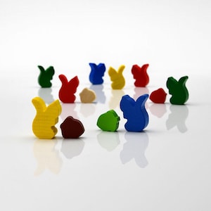 May include: A collection of colorful wooden squirrel-shaped game pieces and acorn-shaped tokens. The pieces are painted in various colors, including yellow, red, green, and blue. The acorns are brown and tan. The pieces are arranged on a white surface.