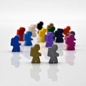 Op de afbeelding: Een verzameling kleine, houten spelstukken in diverse kleuren, waaronder blauw, rood, geel en paars. De stukken zijn gevormd als mensen met rugzakken, op een witte ondergrond. De focus ligt op de diverse kleuren en het uniforme ontwerp.