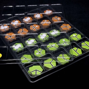 May include: A clear plastic storage tray with a lid, filled with hexagonal tiles in shades of green, orange, and white. The lid has a yellow sticker that says "Open Here".