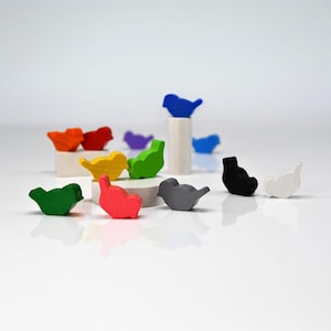 Puede incluir: Una colección de coloridas figuras de pájaros de madera en varios tonos, incluyendo rojo, naranja, amarillo, verde, azul, gris, negro y blanco. Algunos pájaros están posados en pequeñas plataformas blancas. El fondo es una superficie blanca y limpia.