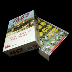 May include: A board game box with the title "1862 Railway Mania in the Eastern Counties" and a vintage illustration of a train station. The box is open, revealing a tray of game pieces.