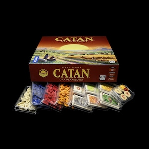 Könnte beinhalten: Ein Brettspielkarton mit dem Titel "CATAN" und einer Landschaftsillustration. Darunter befinden sich transparente Kunststoffschalen mit Spielsteinen in verschiedenen Farben, darunter Beige, Blau, Rot und Orange. Das Spiel ist auf Polnisch.