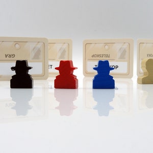 May include: Four wooden game pieces with hats, each representing a different character. The pieces are black, red, blue, and beige. Each piece is in front of a card with a name written on it: "GRA", "ET", "TELESKOP", and "FOKA".