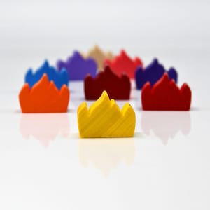 Puede incluir: Una colección de piezas de juego de madera con forma de corona de colores brillantes. Las coronas son de varios colores, incluyendo amarillo, naranja, rojo, azul y morado. La corona amarilla está enfocada, con otras borrosas en el fondo. Estas piezas son probablemente para un juego de mesa.