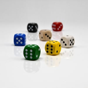 May include: A collection of seven six-sided dice in various colors including green, blue, black, natural wood, yellow, red, and white. Each die features standard pips arranged in numerical patterns from one to six, with contrasting colors for the dots.