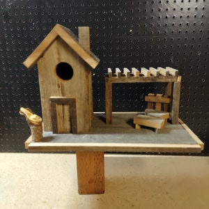 May include: A wooden birdhouse with a small porch and a roof with a chimney. The birdhouse is on a wooden platform with a single wooden post.