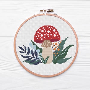May include: A red and white mushroom with white spots, surrounded by green and blue leaves, is embroidered on a white canvas in a wooden embroidery hoop.