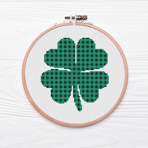 Può includere: Ricamo a punto croce di un quadrifoglio in un telaio di legno. Il quadrifoglio è verde con un motivo a quadri neri. Lo sfondo è bianco.