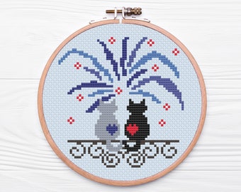 May include: A cross-stitch embroidery hoop with a blue and white firework design and two cats sitting under the fireworks. The cats are gray and black and have red hearts on their chests.