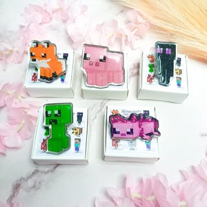 Peut inclure: Cinq broches de personnages Minecraft, dont un renard, un cochon, un enderman, un creeper et un autre cochon, présentées sur de petites boîtes blanches. Les broches sont colorées et détaillées, avec une finition brillante. Le fond présente des décorations florales roses.