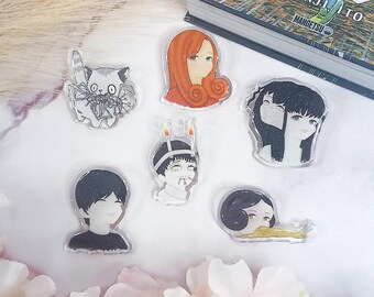 May include: A set of six clear acrylic charms featuring characters from the Junji Ito manga series. The characters include a girl with long red hair, a girl with black hair, a boy with black hair, a boy with a candle on his head, a girl with a snail, and a creature with a spider.