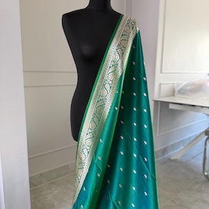 May include: A teal and green silk scarf draped over a black mannequin. The scarf has a detailed silver and gold border with an intricate floral pattern. Small, silver-colored dots are scattered across the fabric.