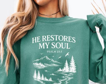 May include: A teal green sweatshirt with the text "HE RESTORES MY SOUL" and "PSALM 23:3" in white. The graphic features a mountain and lake scene with trees and birds. The model is wearing denim shorts.