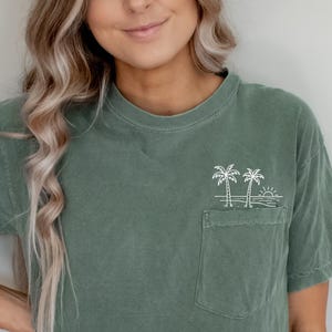 K&ouml;nnte beinhalten: Olivgr&uuml;nes kurz&auml;rmeliges T-Shirt mit einer Brusttasche. Die Tasche hat ein wei&szlig;es gesticktes Design mit zwei Palmen und einer Sonne. Das Shirt wird mit hellblauen Jeans getragen.