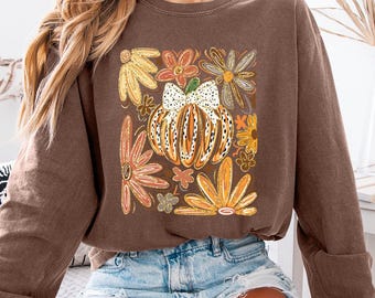 May include: A brown long-sleeved sweatshirt with a fall-themed graphic. The graphic features a pumpkin with a bow, surrounded by orange, yellow, and brown flowers. The sweatshirt is worn with denim shorts.