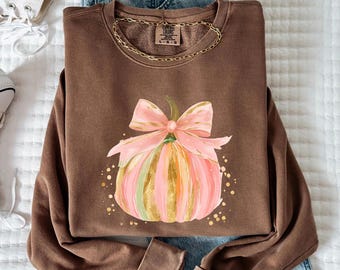 May include: A brown sweatshirt with a pink and gold pumpkin design and a pink bow. The sweatshirt is paired with blue jeans and white high-top sneakers. A gold chain necklace is visible.