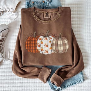 May include: A brown sweatshirt featuring three pumpkin designs in orange, floral, and tan plaid patterns. The sweatshirt is styled with light-wash denim jeans and white high-top trainers. A gold chain necklace is on the sweatshirt.