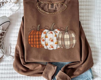 May include: A brown sweatshirt with three pumpkin designs in orange plaid, floral, and tan plaid patterns. The sweatshirt is paired with light-wash denim jeans and white high-top sneakers. A gold chain necklace is draped on the sweatshirt.
