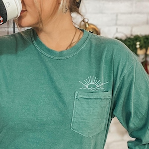 May include: A teal long-sleeve t-shirt with a white pocket and a sunrise graphic. The shirt has a relaxed fit and is made of a soft, comfortable fabric.