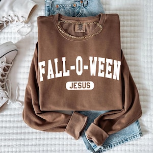 May include: A brown sweatshirt with the text "FALL-O-WEEN JESUS" in white, paired with light blue denim jeans and white high-top sneakers. A gold chain necklace is draped on the sweatshirt. The items are arranged on a white quilted surface.