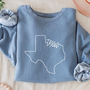 May include: A light blue sweatshirt with the outline of the state of Texas in white, with the word "Texas" in a cursive font. The sleeves are cuffed. A hat and decorative items are in the background.