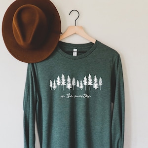 May include: A dark green long-sleeved shirt with a white graphic of a row of trees and the text "on the mountain".