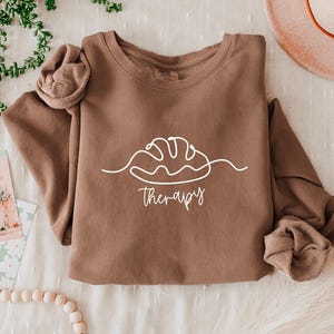 May include: A brown long-sleeved sweatshirt with a white line drawing of a loaf of bread and the word "therapy" in a script font. The sleeves are cuffed. A hat and decorative items are in the background.