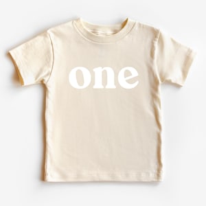 May include: A cream-colored t-shirt with the word "one" printed in white block letters. The short-sleeved shirt is made of a soft material and is suitable for a child's first birthday celebration.