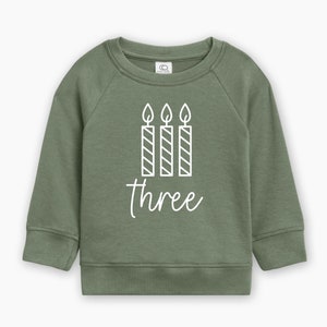 May include: Green sweatshirt with a white graphic of three birthday candles and the word "three" on it.