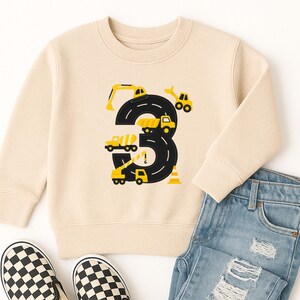 May include: A beige sweatshirt with a black number 3 design featuring yellow construction vehicles. The outfit includes light blue ripped jeans and black and white checkered slip-on shoes. A fun outfit for a child's birthday.
