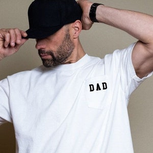May include: A white t-shirt with a black pocket that says "DAD" on it. The person is wearing a black baseball cap.