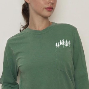 May include: A long-sleeved, forest green shirt with a small pocket on the chest. The pocket features a white graphic of several pine trees. The shirt is paired with olive green shorts. The shirt is made of a soft, comfortable material.