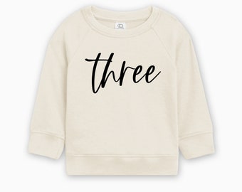 May include: A cream colored sweatshirt with the word "three" written in black cursive lettering.